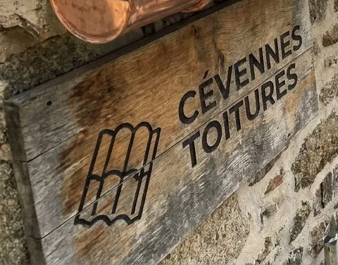 Enseigne en bois Cévennes Toitures sur un mur en pierre sous une gouttière en cuivre, près d'Alès